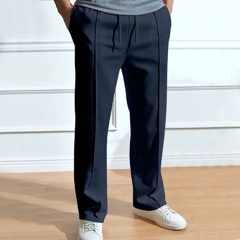 Model wearing the trousers in navy with hands in pockets, showing front pintuck creases and drawstring waist, with white trainers. 1
