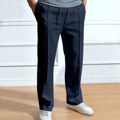 Model wearing the trousers in navy with hands in pockets, showing front pintuck creases and drawstring waist, with white trainers. 1