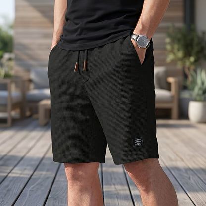 Model wearing black waffle-textured shorts with drawstring and small woven label, hand in pocket on a wooden deck 3