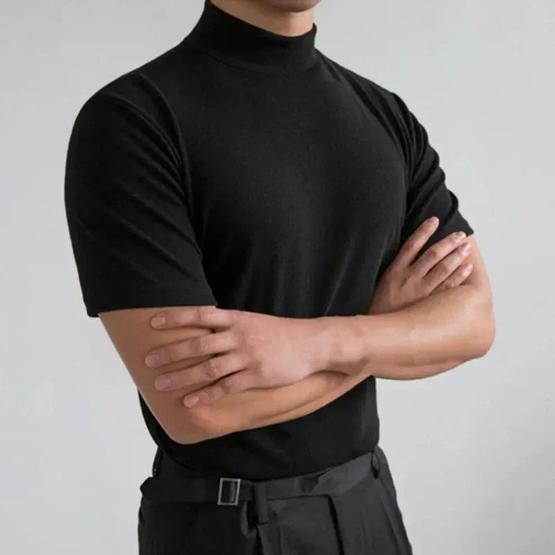 Same black mock-neck top worn with arms crossed against a pale grey wall, showing the fitted shoulders and sleeves 2