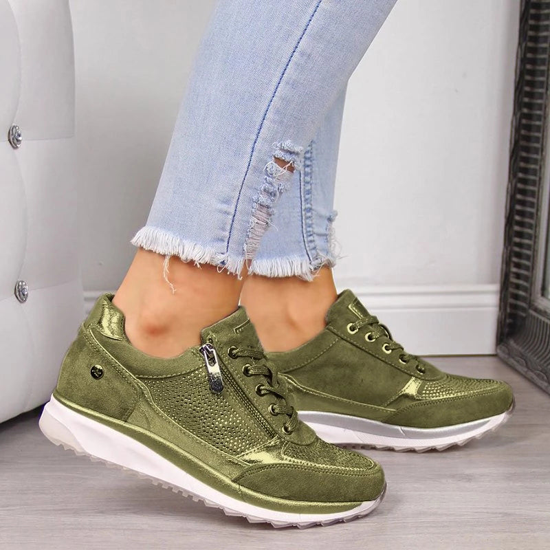 Olive-green version of the trainers with metallic trim and side zip, shown on feet beside a white padded wall. 4