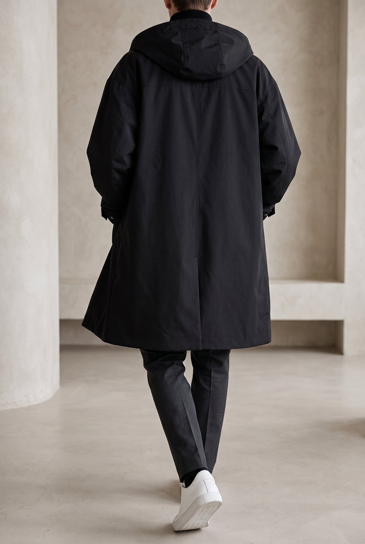 Back view of the black hooded coat on a model walking, showing hood and loose, long cut 7