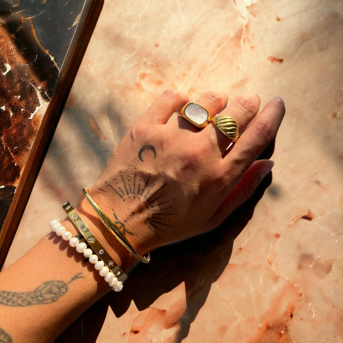Tattooed hand and wrist wearing the gold cuff layered with a crystal-studded bangle, a pearl bracelet and two gold rings, resting on pink marble. 4
