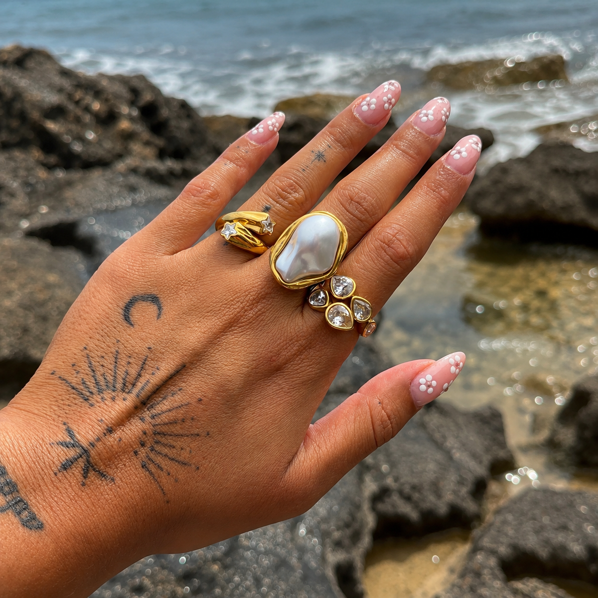 Hand held up against rocks and sea, wearing the pear-crystal ring beside a large gold-framed pearl ring and a star-set ring. 5