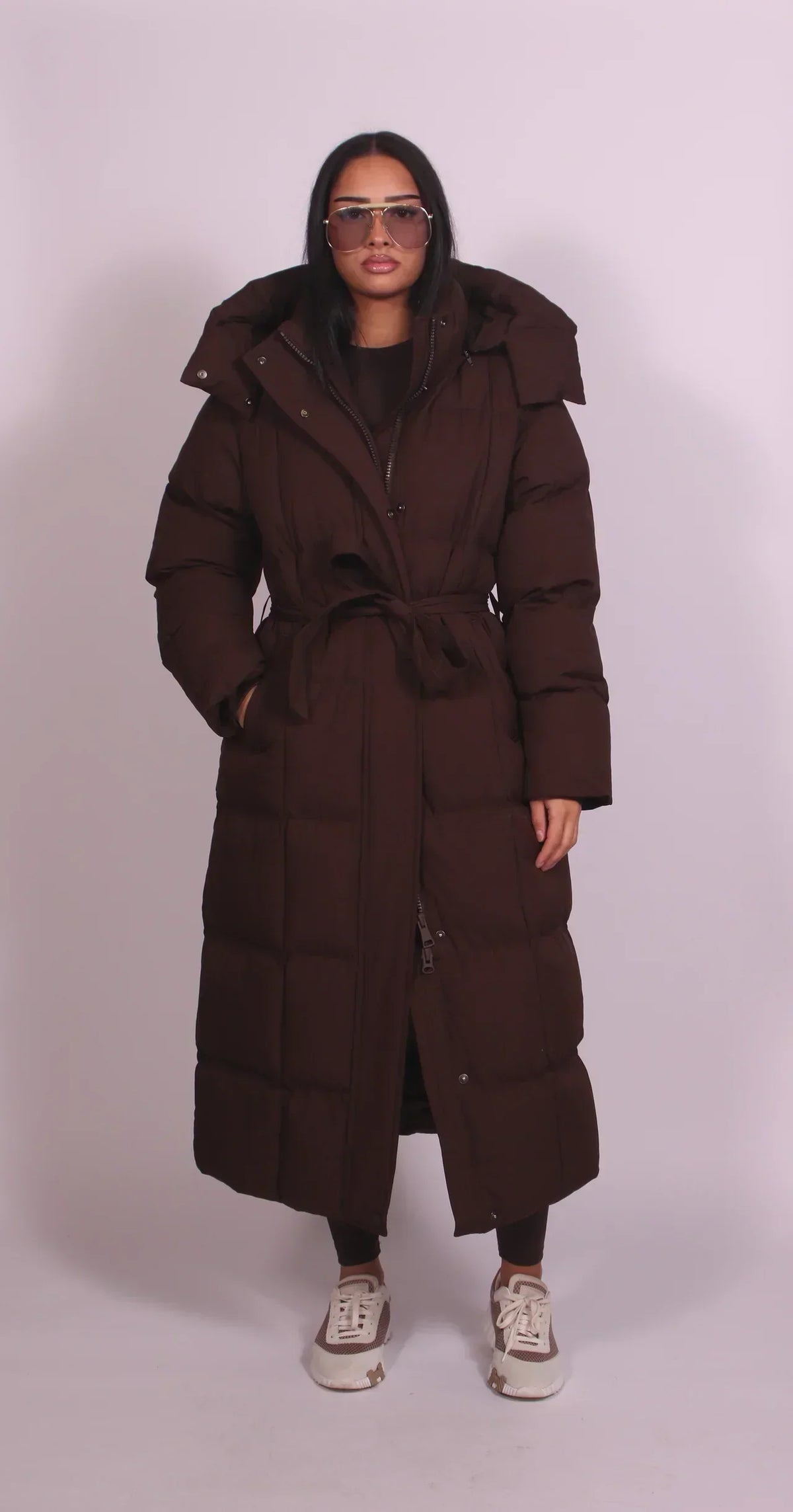 Front view of the dark brown quilted coat, belted at the waist with the hood up, worn by a model 1