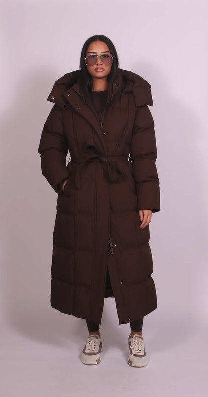 Front view of the dark brown quilted coat, belted at the waist with the hood up, worn by a model 1