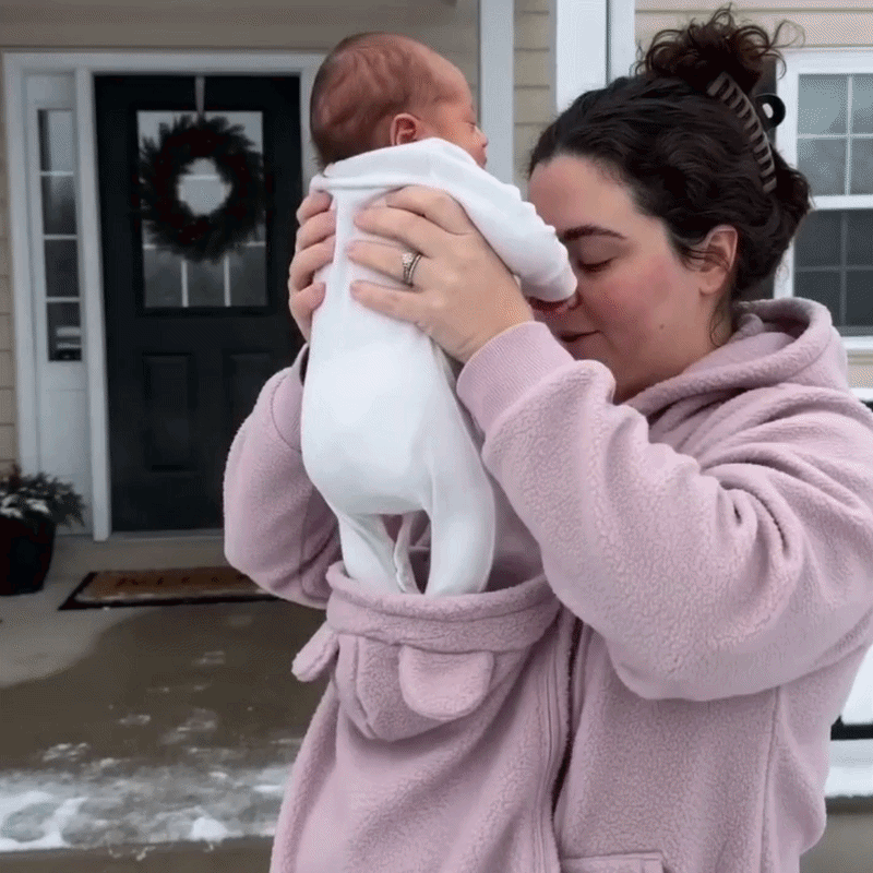 Woman in the pink fleece hoodie lifting a baby in white out of the front pouch outside a front door. 11