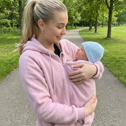 Woman in a pink fleece hoodie on a park path, gazing at a baby in a blue knitted hat in the carrier. 4