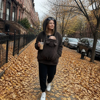 Woman in a dark brown sherpa hoodie with the hood up walking down a leaf-covered street, baby snug in the front panel. 5