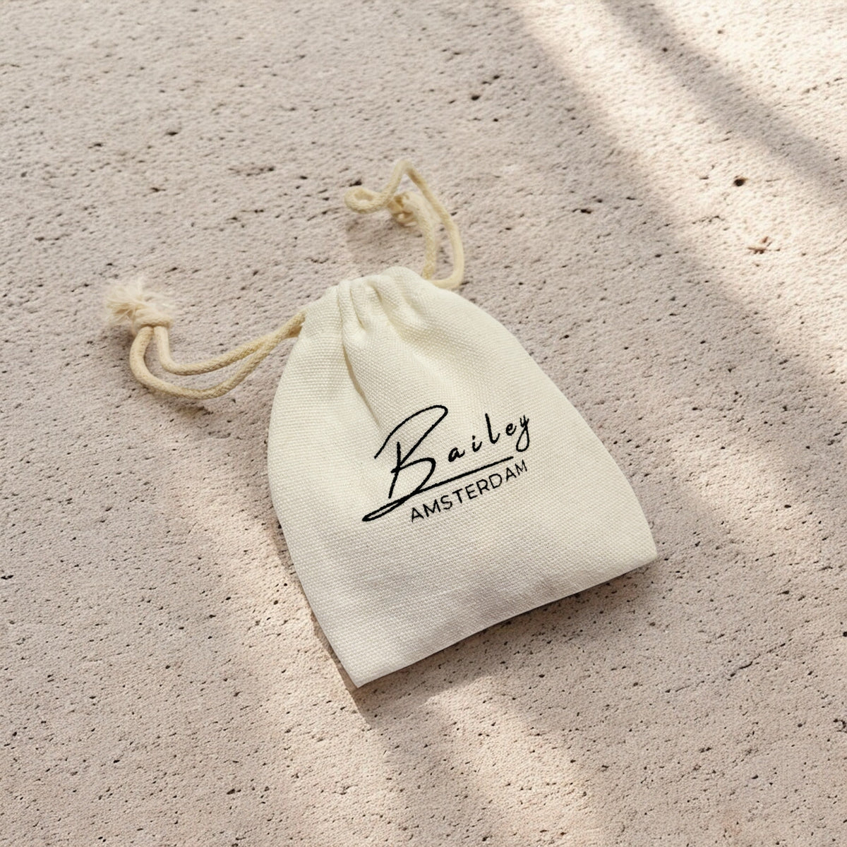 Small cream cotton drawstring pouch with a black script logo, lying on a sandy textured surface in dappled sunlight, used as jewellery packaging. 6