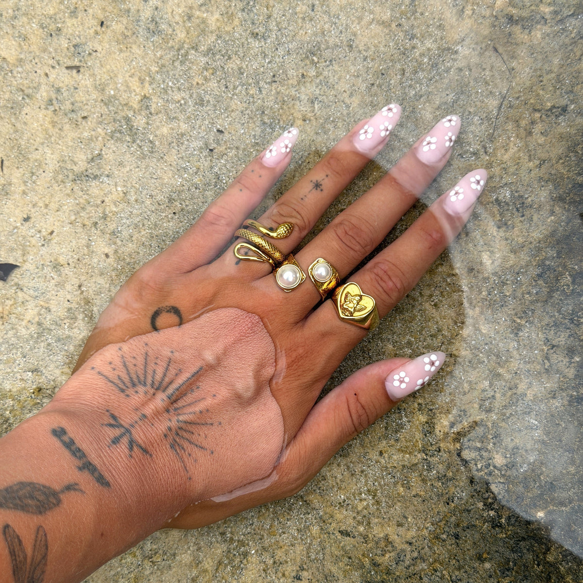 Tattooed hand with pink floral nails wearing the heart cherub ring beside a snake ring and pearl rings 4