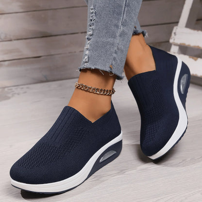 Navy blue knit slip-on trainers with white soles and air cushion, worn on a model's feet beside a wooden step. 2