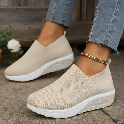 Beige knit slip-on trainers with white soles and clear air cushion, worn with cuffed jeans on a stone path. 3