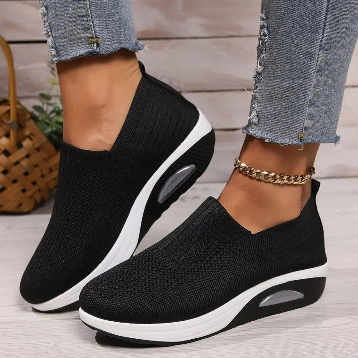 Black knit slip-on trainers with white platform soles and visible air cushion, worn with frayed jeans and a gold anklet. 1