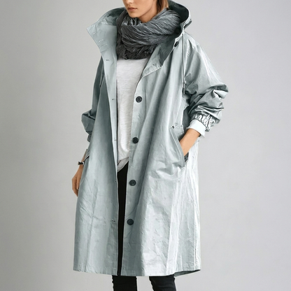 Model wearing the long hooded coat in a pale silvery blue-grey with a slightly glossy finish, hand in pocket, grey scarf. 5