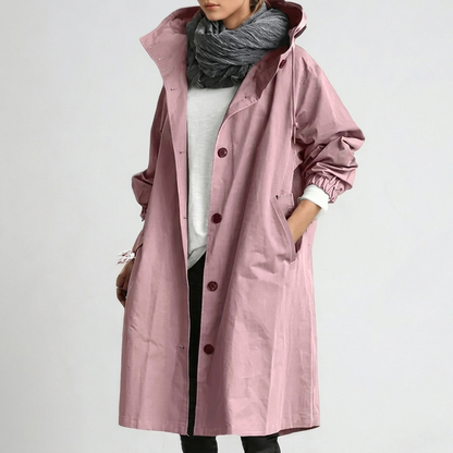 Model wearing the long hooded button-up coat in dusky pink, one hand in the pocket, with a grey scarf and white top. 1