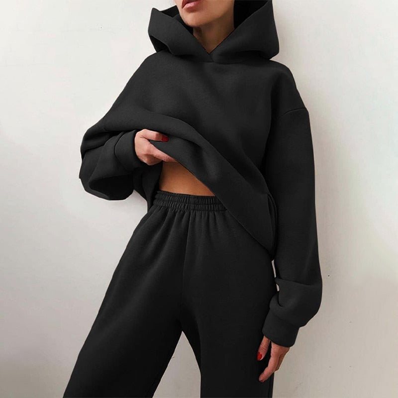Model leaning against a wall in the matching oversized hoodie and elastic-waist joggers in black, one hand lifting the hem. 2