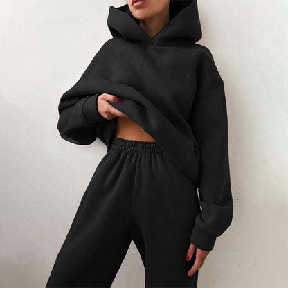 Model leaning against a wall in the matching oversized hoodie and elastic-waist joggers in black, one hand lifting the hem. 2