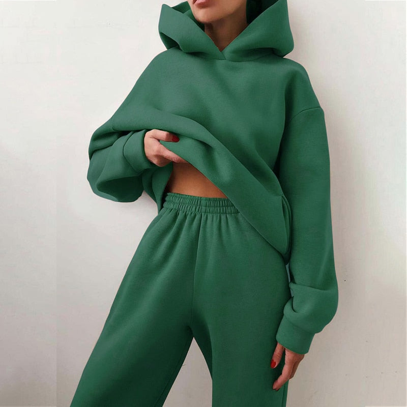 Model in the deep green hoodie and matching elastic-waist joggers, holding the hem of the loose-fitting top. 9
