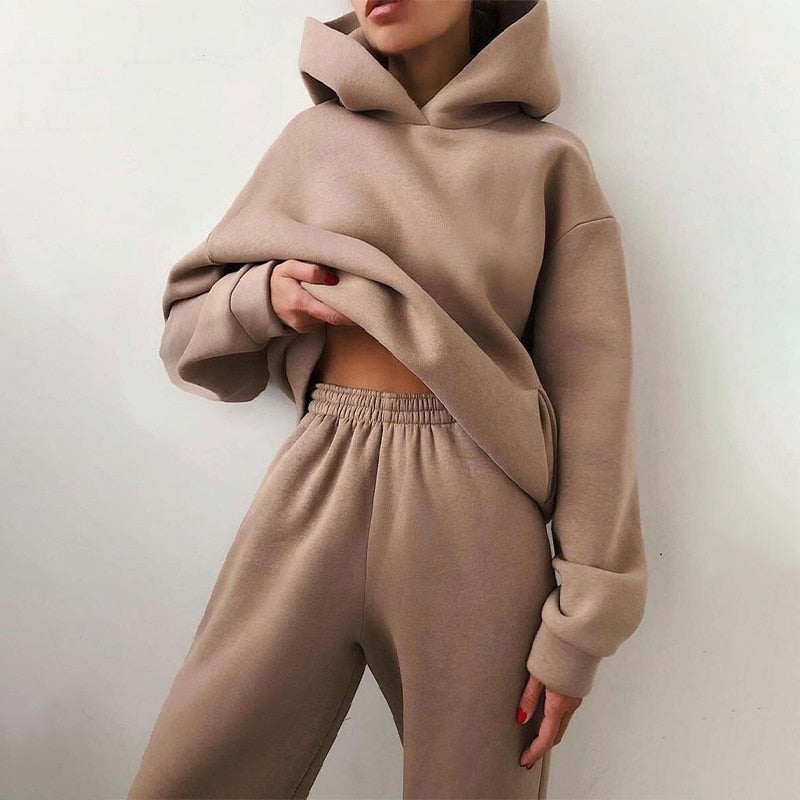 Model leaning against a wall in the oversized hoodie and elastic-waist joggers, in a light taupe-brown, one hand lifting the hem. 1