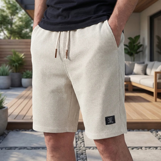 Close three-quarter view of waffle-textured cream shorts with drawstring and hand in pocket, worn on a sunny patio 2