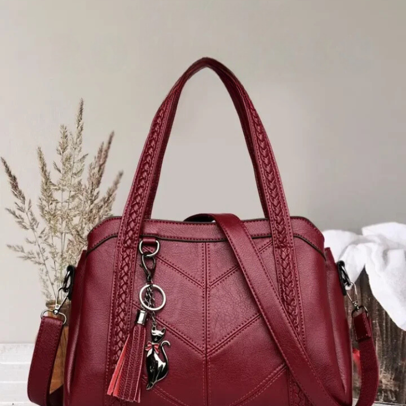 Burgundy handbag with braided handles, tassel and cat charm, styled against a pale wall beside dried grasses and a white cloth. 2