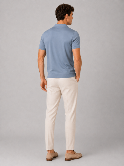 Back view of the dusty blue polo on a standing model, showing the plain back, short sleeves and hem, with cream trousers and loafers. 19
