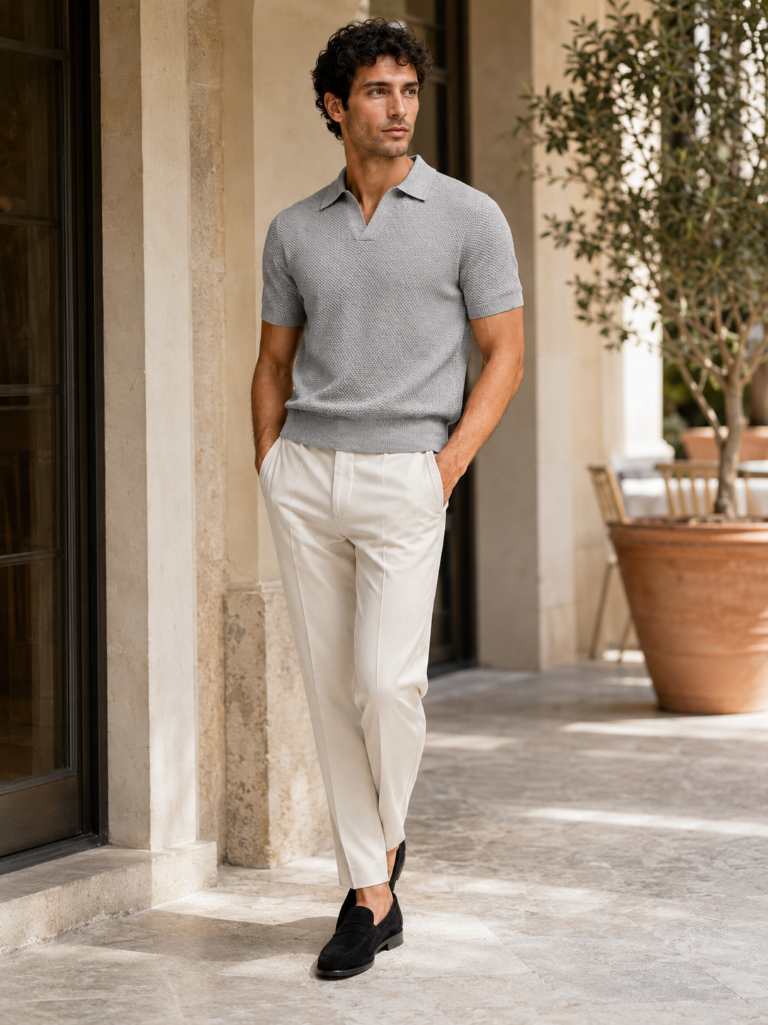 Model in grey knit polo and cream trousers standing on a sunlit stone terrace near an olive tree 30