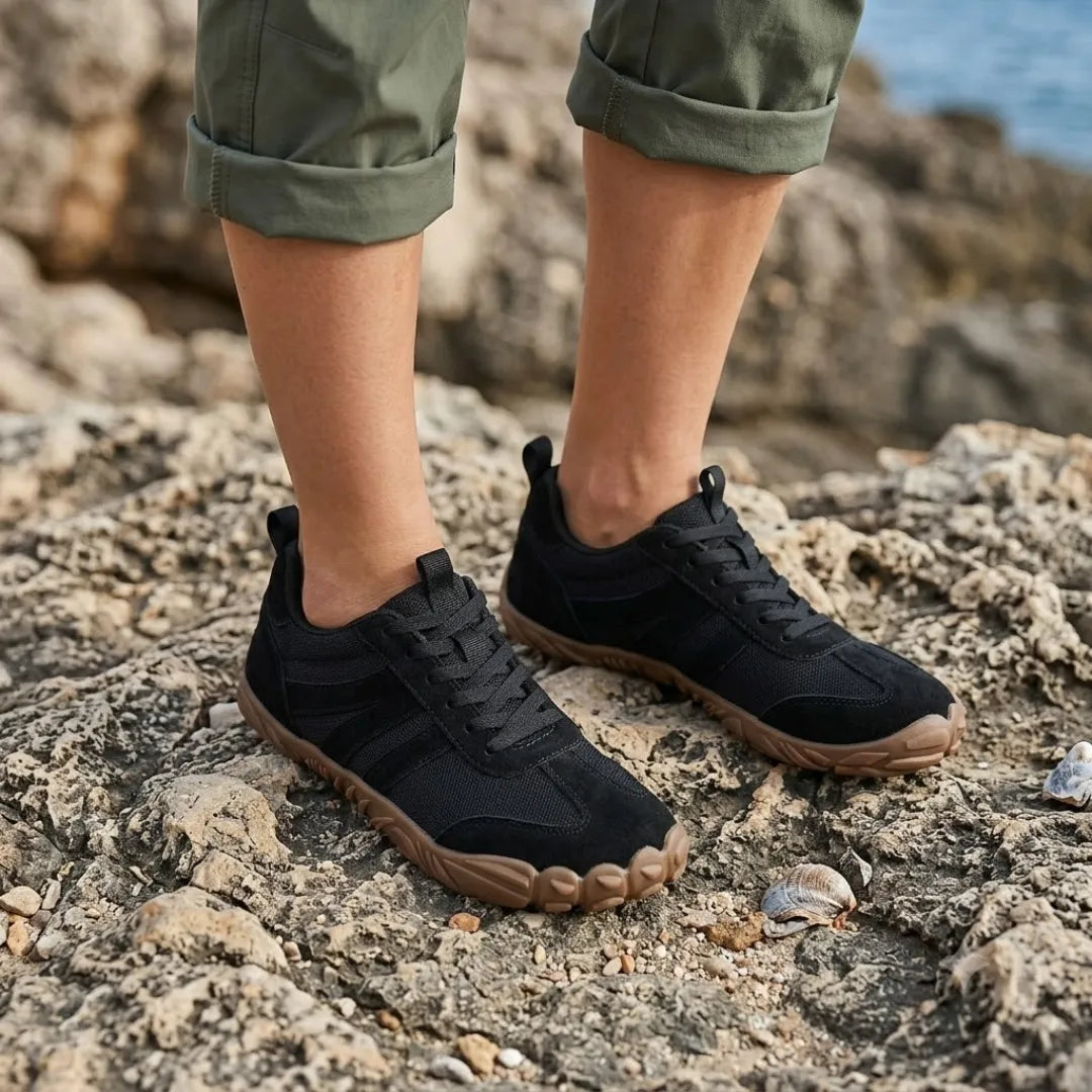 Person wearing the black barefoot shoes with rolled olive trousers, standing on rocky ground near the sea. 20