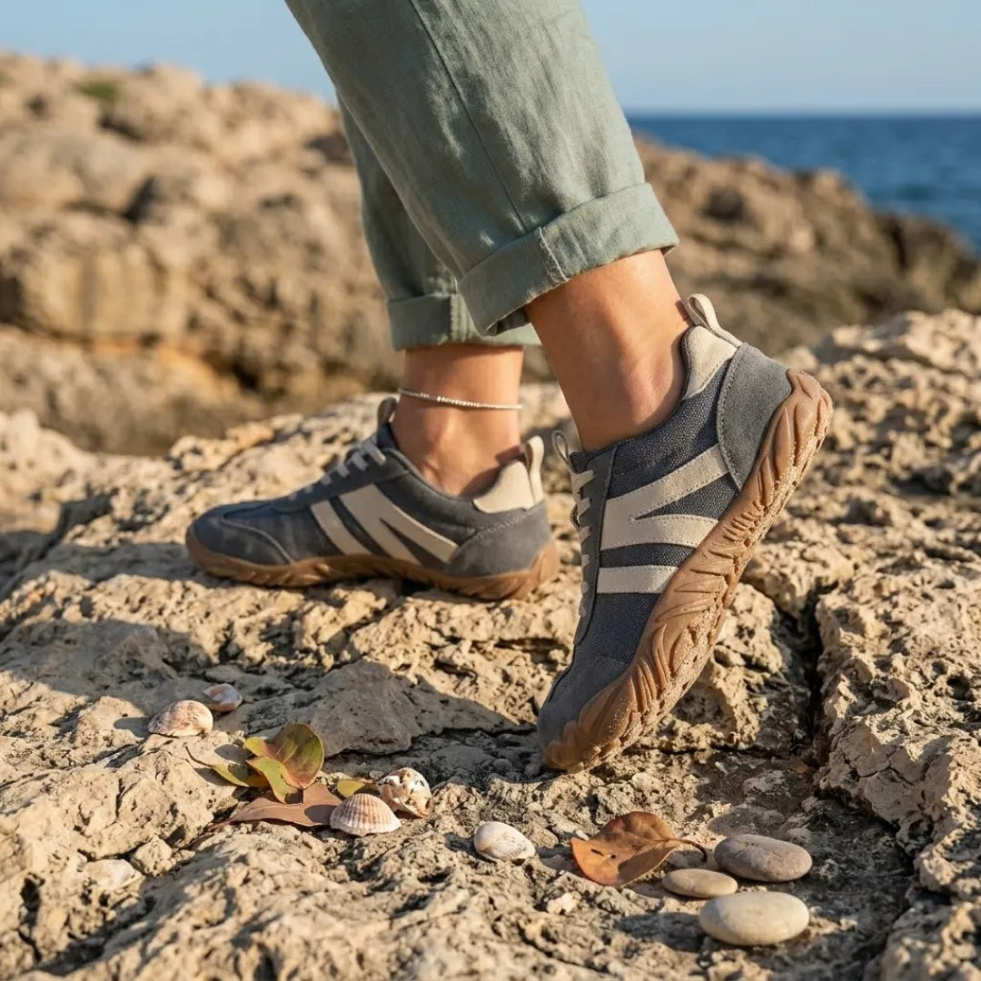 Grey barefoot shoes with cream stripes worn on coastal rocks, with rolled green linen trousers, shells and pebbles nearby. 2