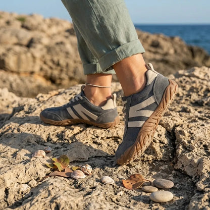 Grey barefoot shoes with cream stripes worn on coastal rocks, with rolled green linen trousers, shells and pebbles nearby. 2