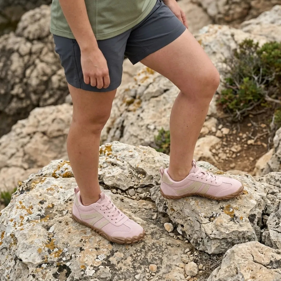 Woman in grey shorts standing on rocky ground wearing the pink barefoot shoes with beige stripes. 11