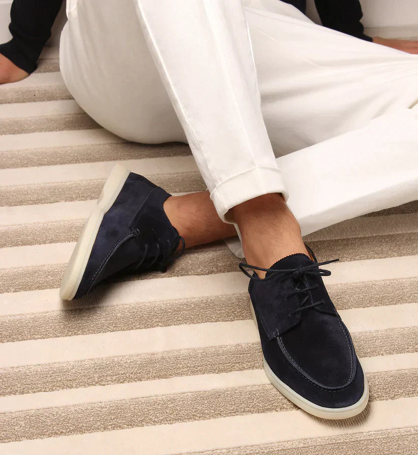 Man seated on a striped rug wearing navy suede lace-up loafers with pale soles, styled with white cuffed trousers. 1