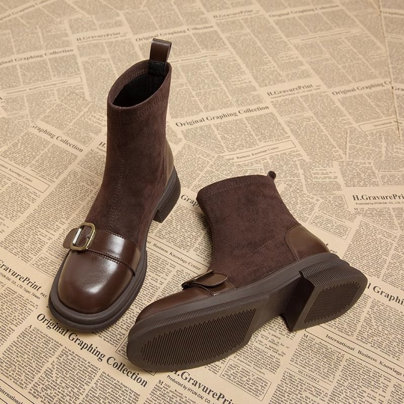Brown suede ankle boots with glossy leather toe caps and brass buckles, one boot lying on its side to show the ridged rubber sole. 1