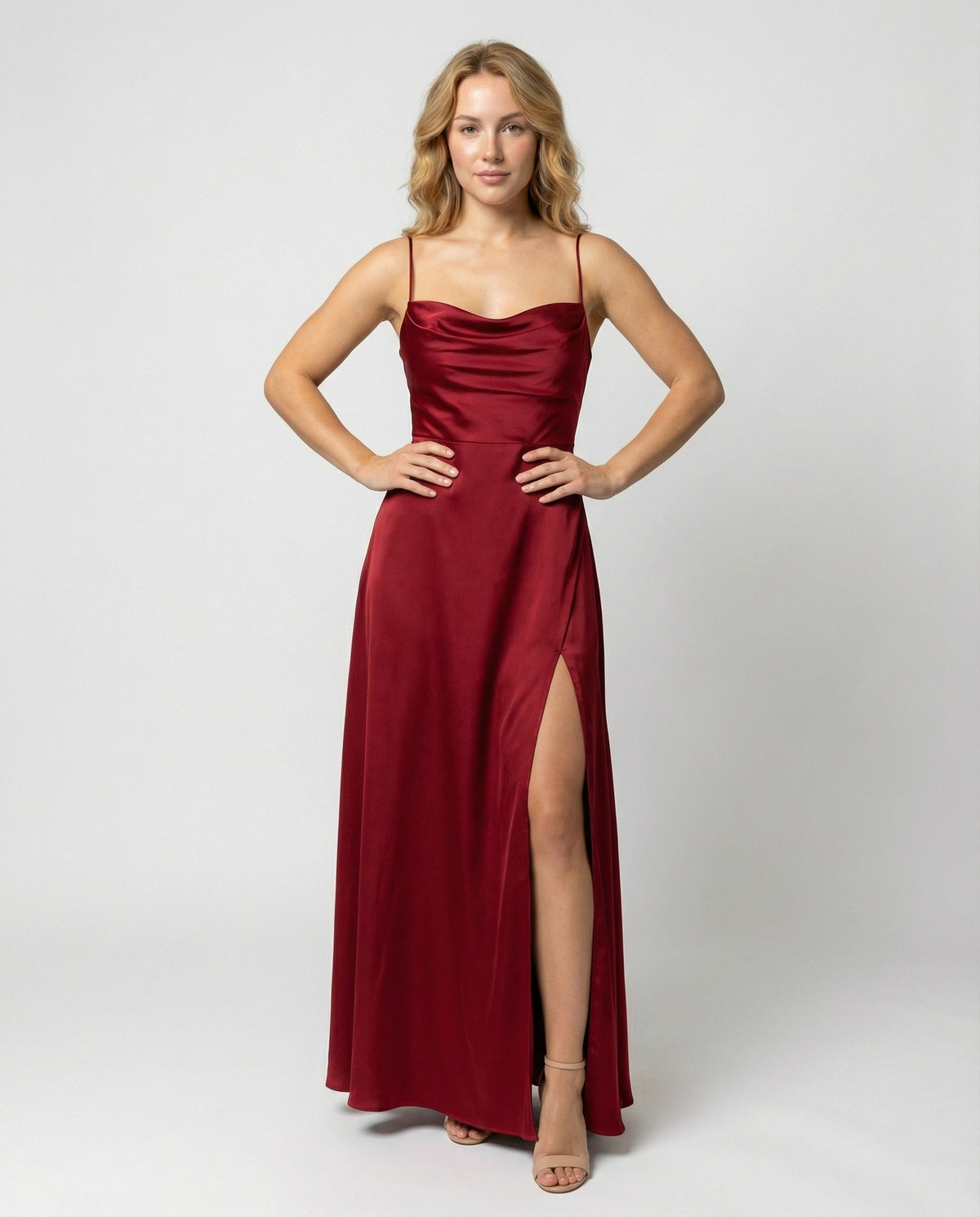 Model with wavy blonde hair stands facing forward, hands on hips, in a burgundy satin cowl-neck dress with thin straps and a high thigh slit. 1