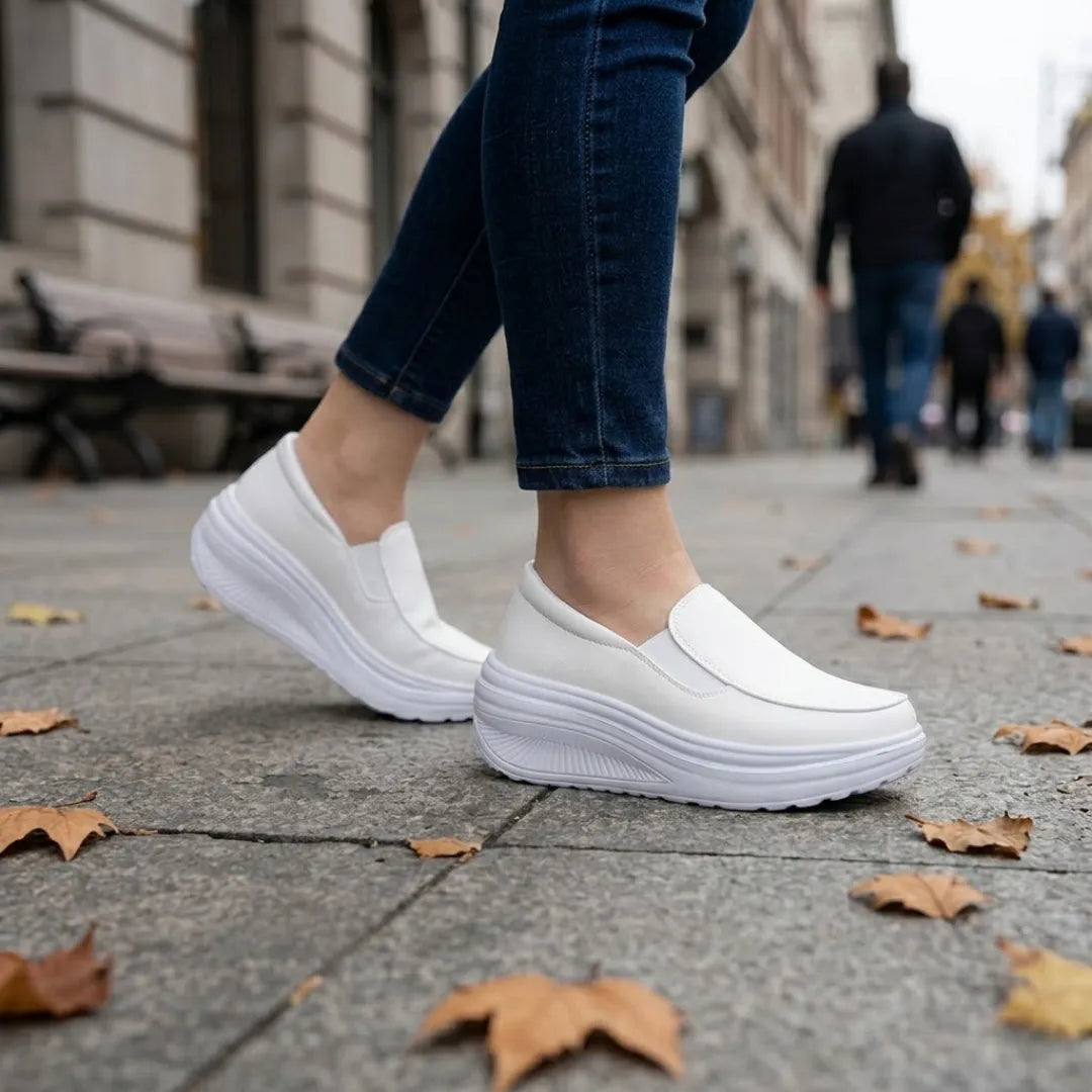 White slip-on shoes worn by a woman in dark jeans, walking on a leaf-strewn pavement 11