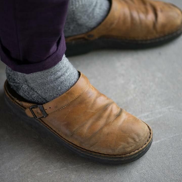 Close-up of a worn tan leather clog on a foot in a grey sock and purple trousers, standing on concrete. 5