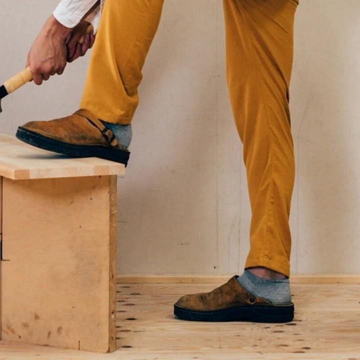 Man in mustard trousers and grey socks wearing tan clogs, one foot raised on a wooden bench while holding a hammer. 7