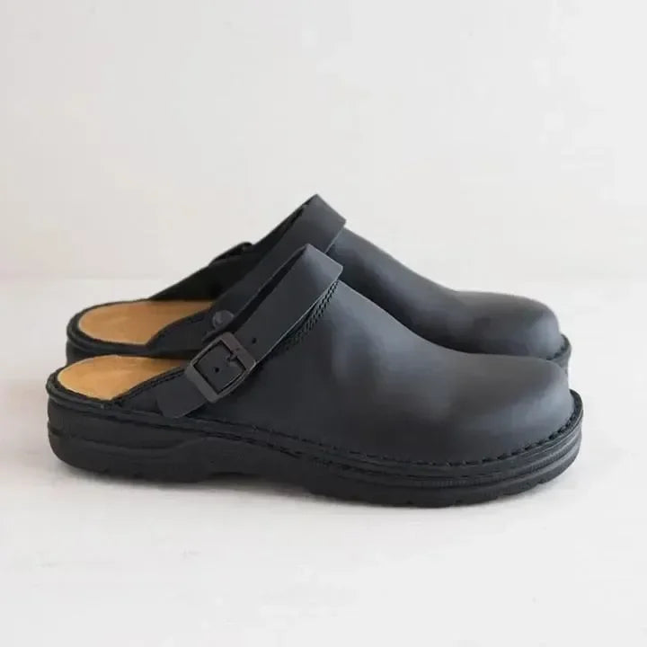 Side view of a pair of black leather mule clogs with an adjustable buckled heel strap, tan footbed and chunky black sole. 9