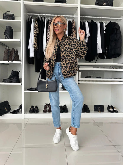 Full-length view of a model in the leopard print bomber jacket, black top, light jeans and white trainers, holding a black shoulder bag. 5