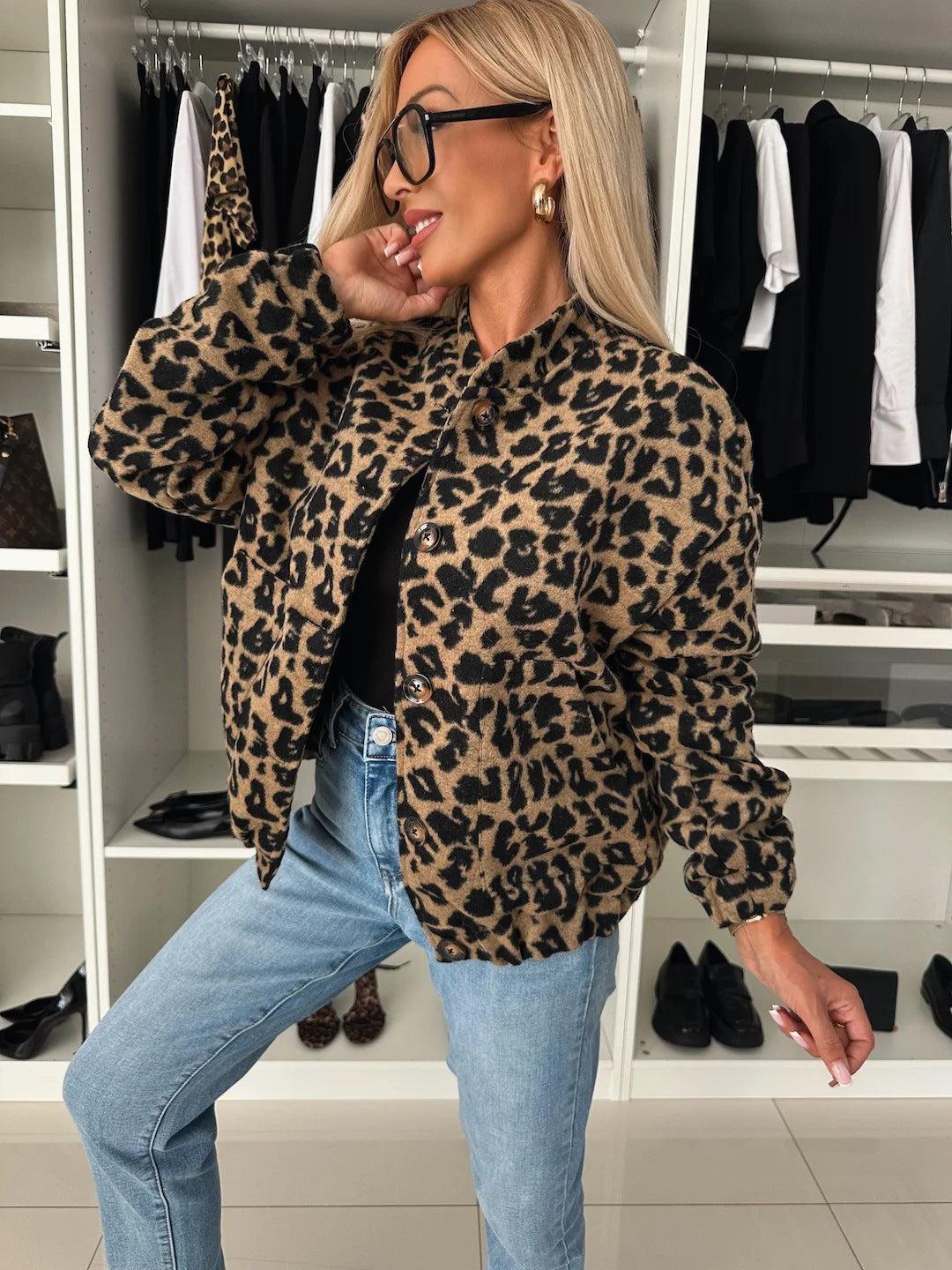 Woman in a wardrobe wearing the buttoned leopard print bomber jacket with blue jeans, her hand raised to her face, seen from the front. 1