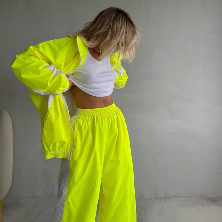 Model in a neon yellow zip jacket and elasticated wide-leg trousers with white trim, lifting a white top against a grey wall. 6
