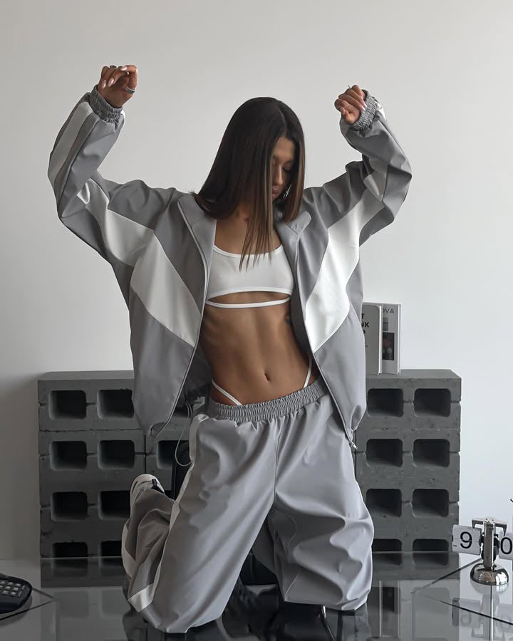Model kneeling with arms raised in a grey windbreaker set with white chevron panels, jacket open over a white bralette. 20