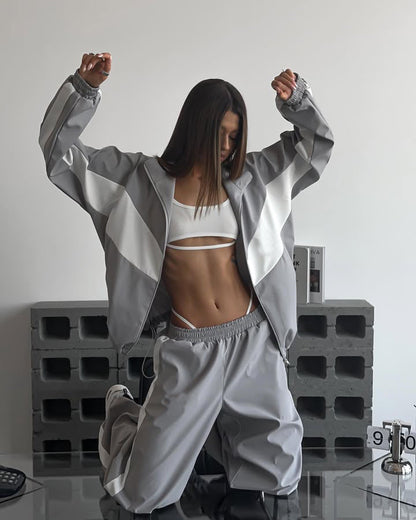 Model kneeling with arms raised in a grey windbreaker set with white chevron panels, jacket open over a white bralette. 20