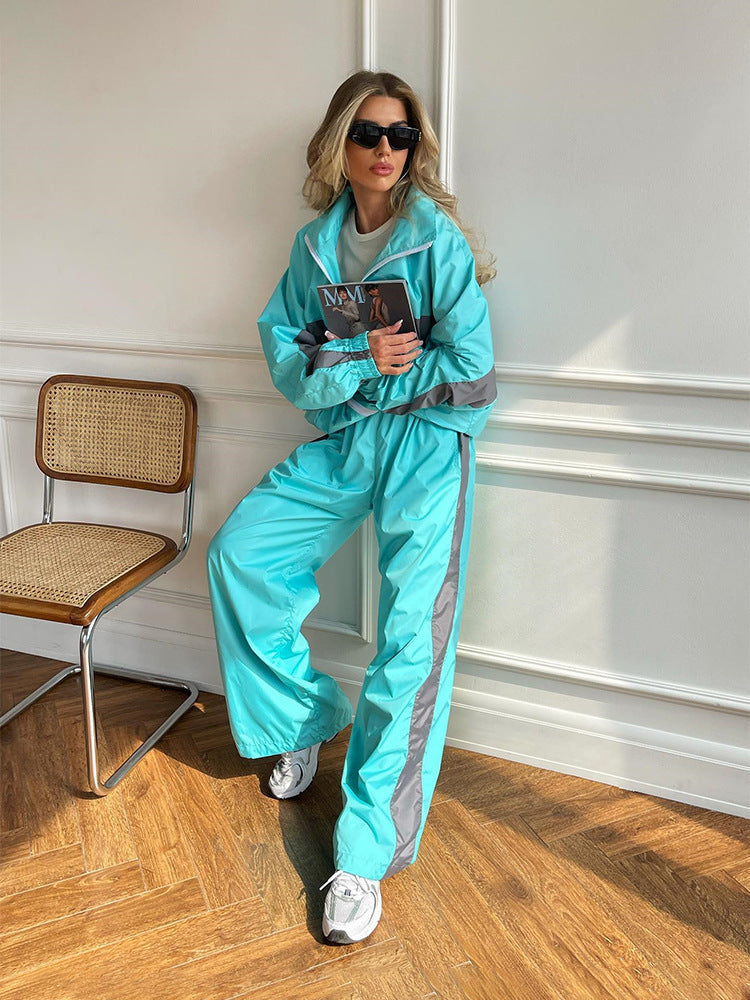 Full-length view of a model leaning on a wall in a turquoise windbreaker set with grey stripes, beside a wooden cane chair. 11