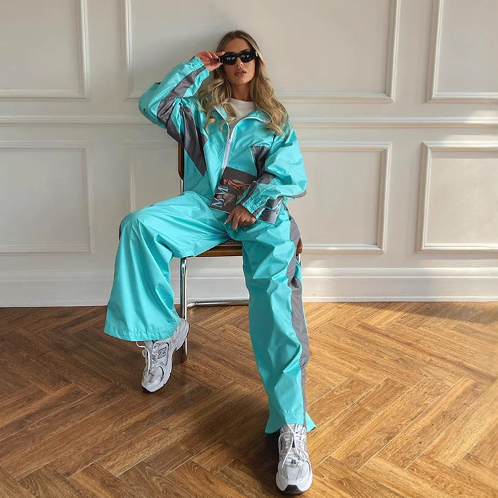 Model sitting on a chair in a turquoise jacket and wide-leg trousers with grey panels, holding a magazine and sunglasses. 4