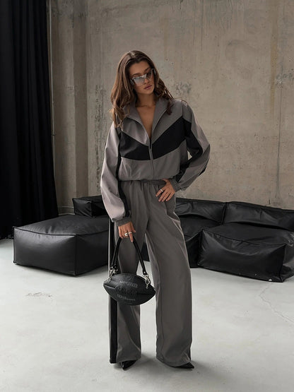 Model standing in the grey set with black chevron panels on the zip-up jacket, wide-leg trousers and sunglasses. 23