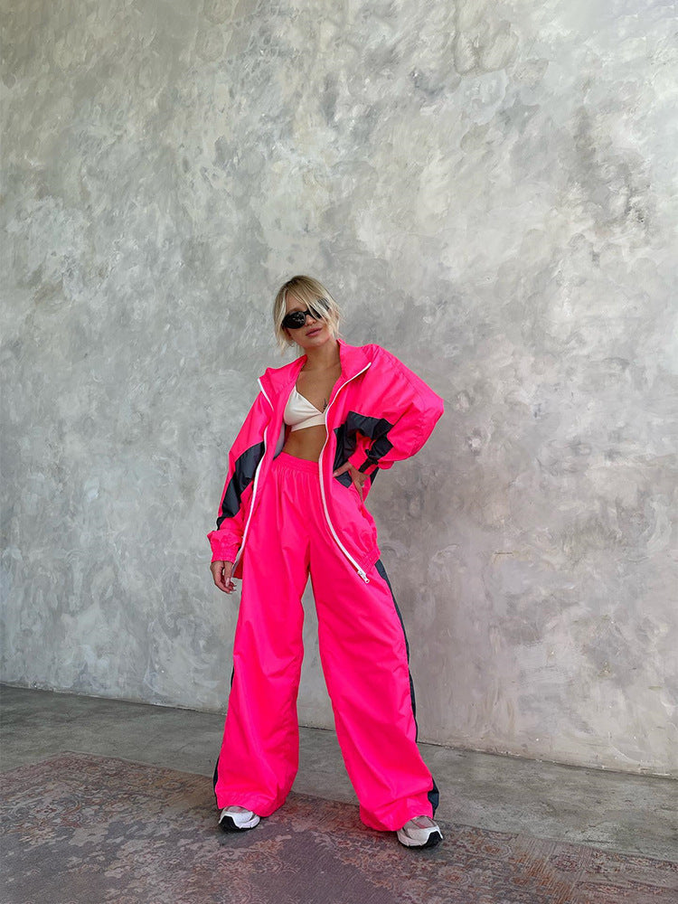 Full-length view of a model in a neon pink windbreaker set with dark grey panels and white zip, standing against a textured wall. 18