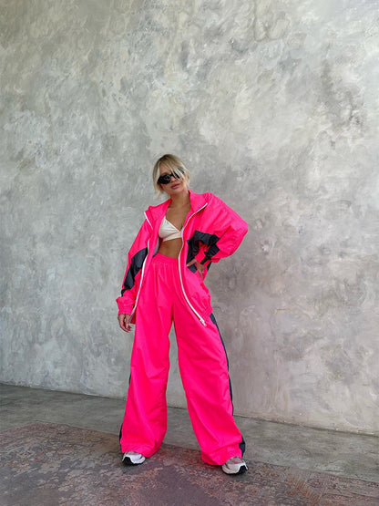 Full-length view of a model in a neon pink windbreaker set with dark grey panels and white zip, standing against a textured wall. 18