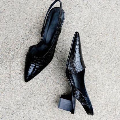 Flat lay of a pair of black crocodile-embossed slingback heels on a concrete floor, showing the upper, strap and chunky heel. 3